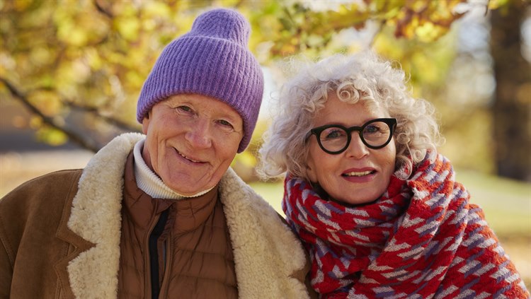 Ett seniorpar står tillsammans med höstfärgade träd bakom sig. De ler mot kameran.