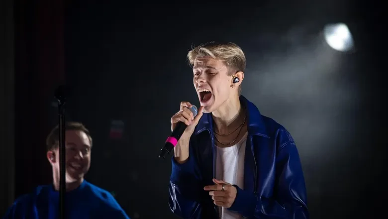 Närbild av en ung blond man i blå kavaj och vit t-shirt som sjunger i en mikrofon